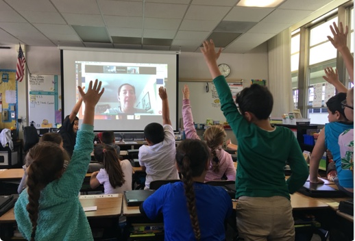 A classroom of students with hands raised looking at a projector, seen from the rear of the room.